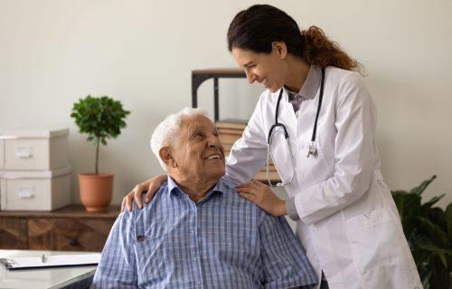 Laughter is the best medicine. Optimistic young lady doc embrace shoulders of laughing old man sitting on chair at clinic office. Doctor joking with aged patient give support motivation on good result